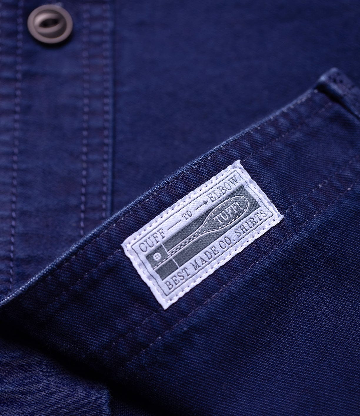 A close-up of the Indigo Moleskin Work Shirt by Best Made Co shows a white stitched "Cuff to Elbow Tuff" label near the pocket, triple-needle stitching, a button, and part of the placket.