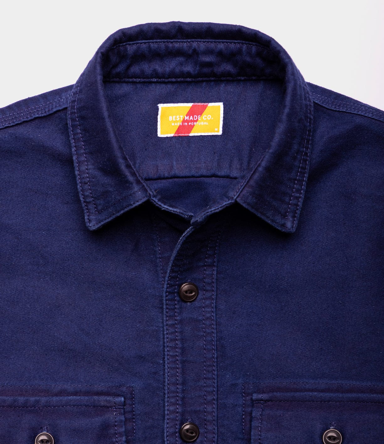 A close-up of the Best Made Co Indigo Moleskin Work Shirt, navy blue with a collar and front pockets. The inside collar label reads “Best Made Co. New York” on a yellow background featuring a red diagonal stripe.