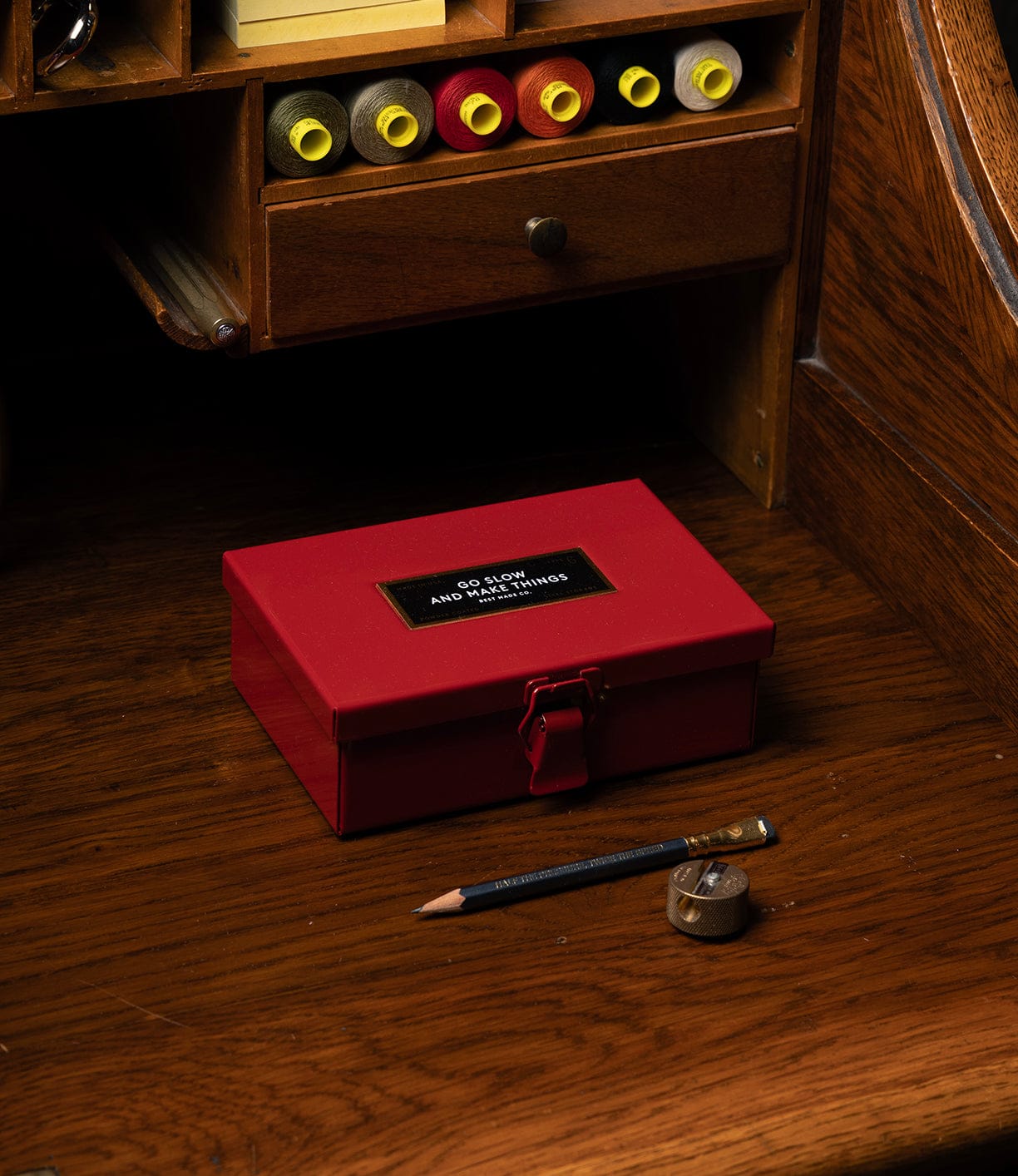 The Best Made Co Type 6 Strongbox, Go Slow Edition, featuring a gold label and clasp, rests on a wooden desk with a pencil and metal sharpener. Above, shelves display yellow and red containers.