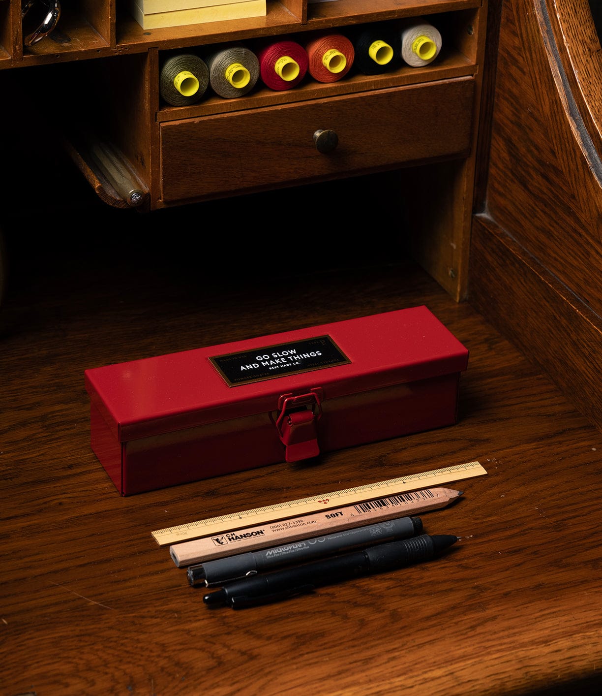 A Best Made Co Type P Strongbox, Go Slow Edition sits on a wooden desk with two pens, a pencil, and a wooden ruler in front. Behind the sturdy box are drawers and shelves holding paper rolls and other stationery supplies.
