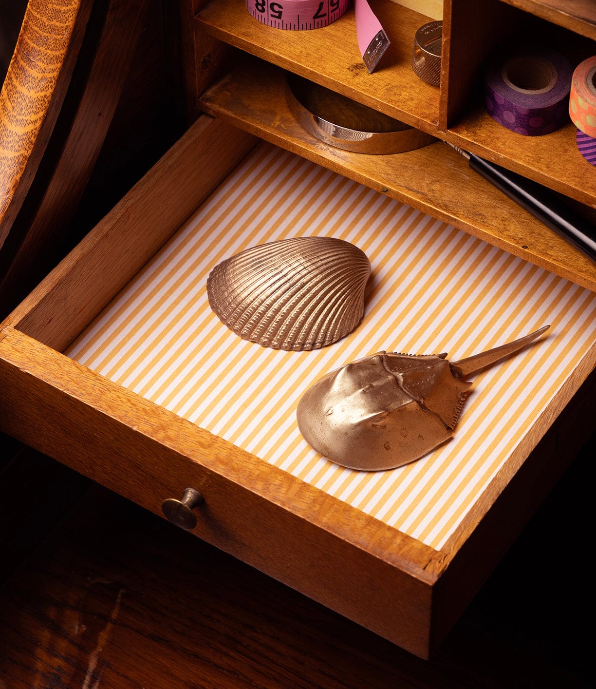 An open wooden drawer lined with yellow and white stripes holds a metallic seashell and the Best Made Co. Horseshoe Crab Opener. Above the drawer are small compartments filled with colorful ribbons and stationery.