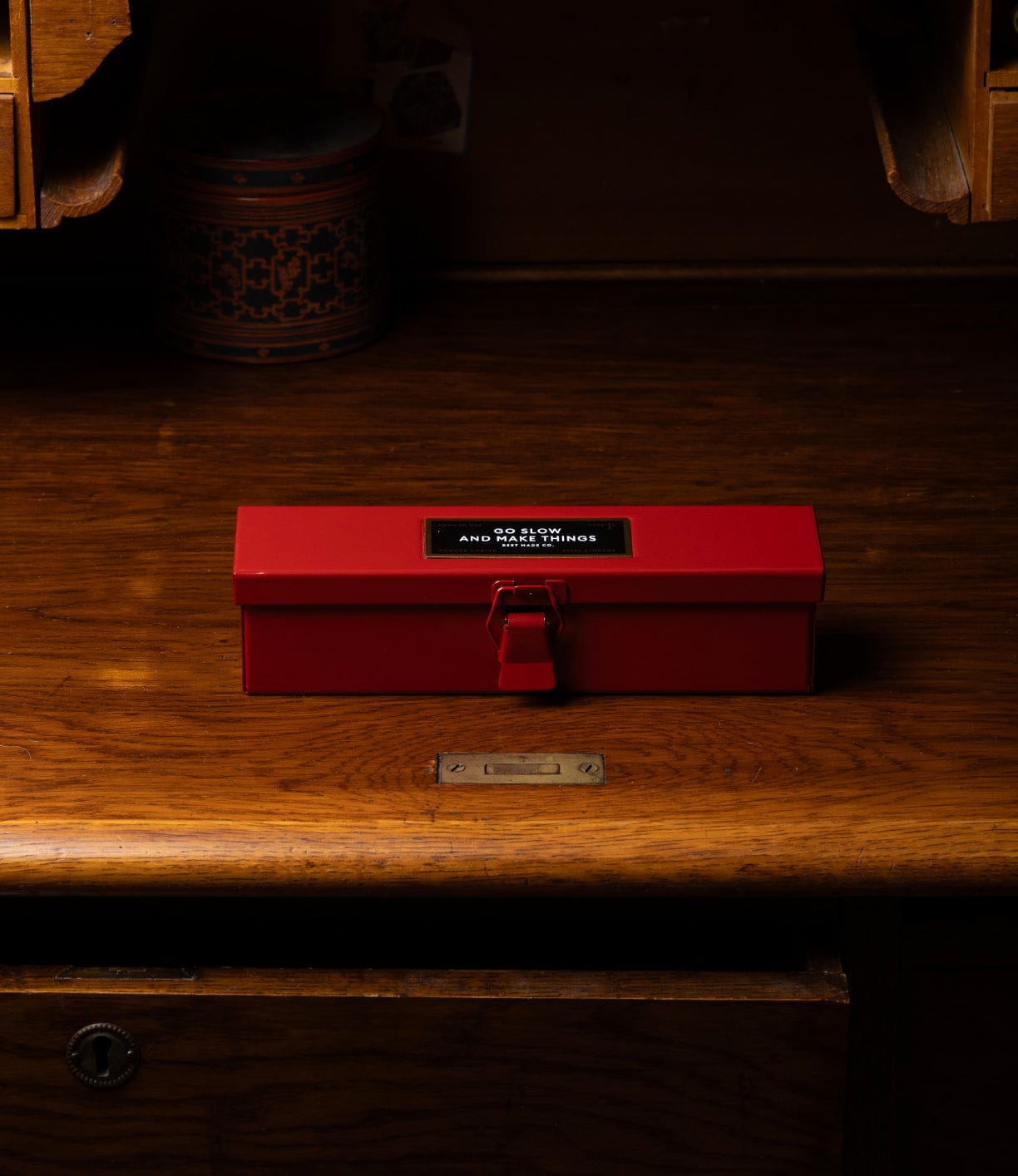 A Type P Strongbox, Go Slow Edition by Best Made Co—a red rectangular steel box with latch and a black label with white text—sits on a wooden desk, the warm lighting creating a vintage ambiance.
