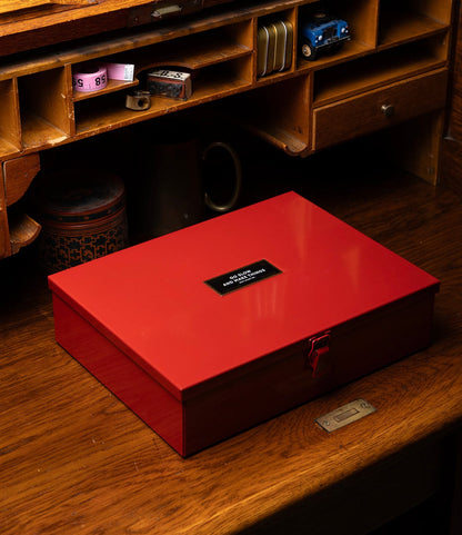 A red Best Made Co. Type 12 Strongbox, Go Slow Edition, labeled "In Case Of Emergency Break Glass," sits on a wooden desk with a vintage organizer, shelves, and small items in the background.