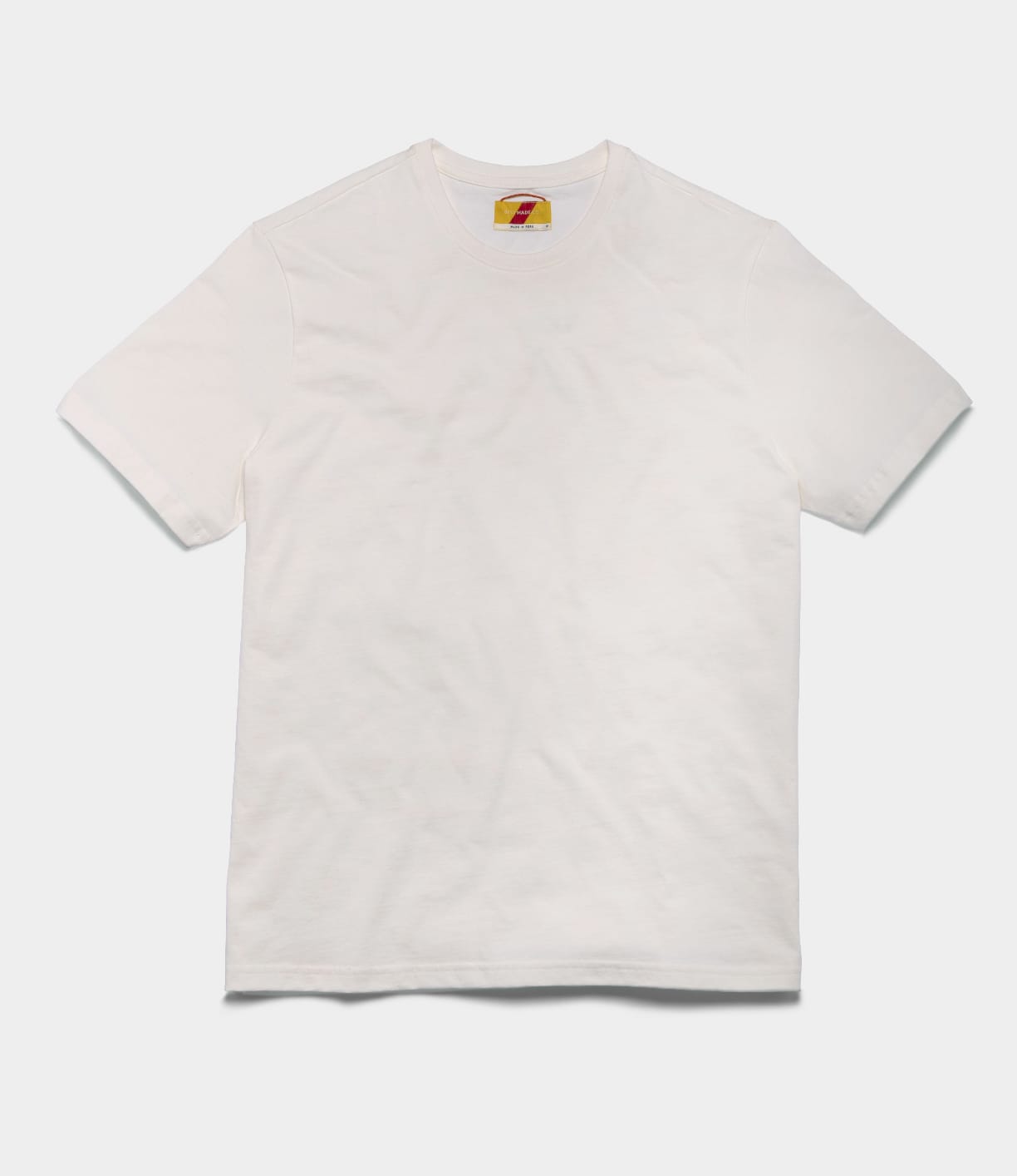 The Best Made Co Standard T-Shirt Undyed, a plain white short-sleeve tee made from Peruvian tangüis cotton, is laid flat on a light background with a small, colorful tag visible inside the neckline.