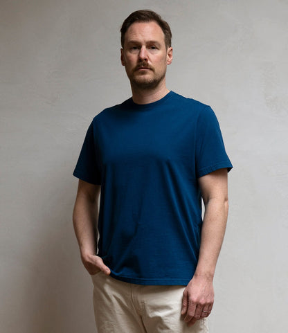 A man with short hair and a mustache stands against a light wall, wearing Best Made Co.'s Standard T-Shirt in Signal Blue and beige pants, one hand in his pocket and a neutral expression.