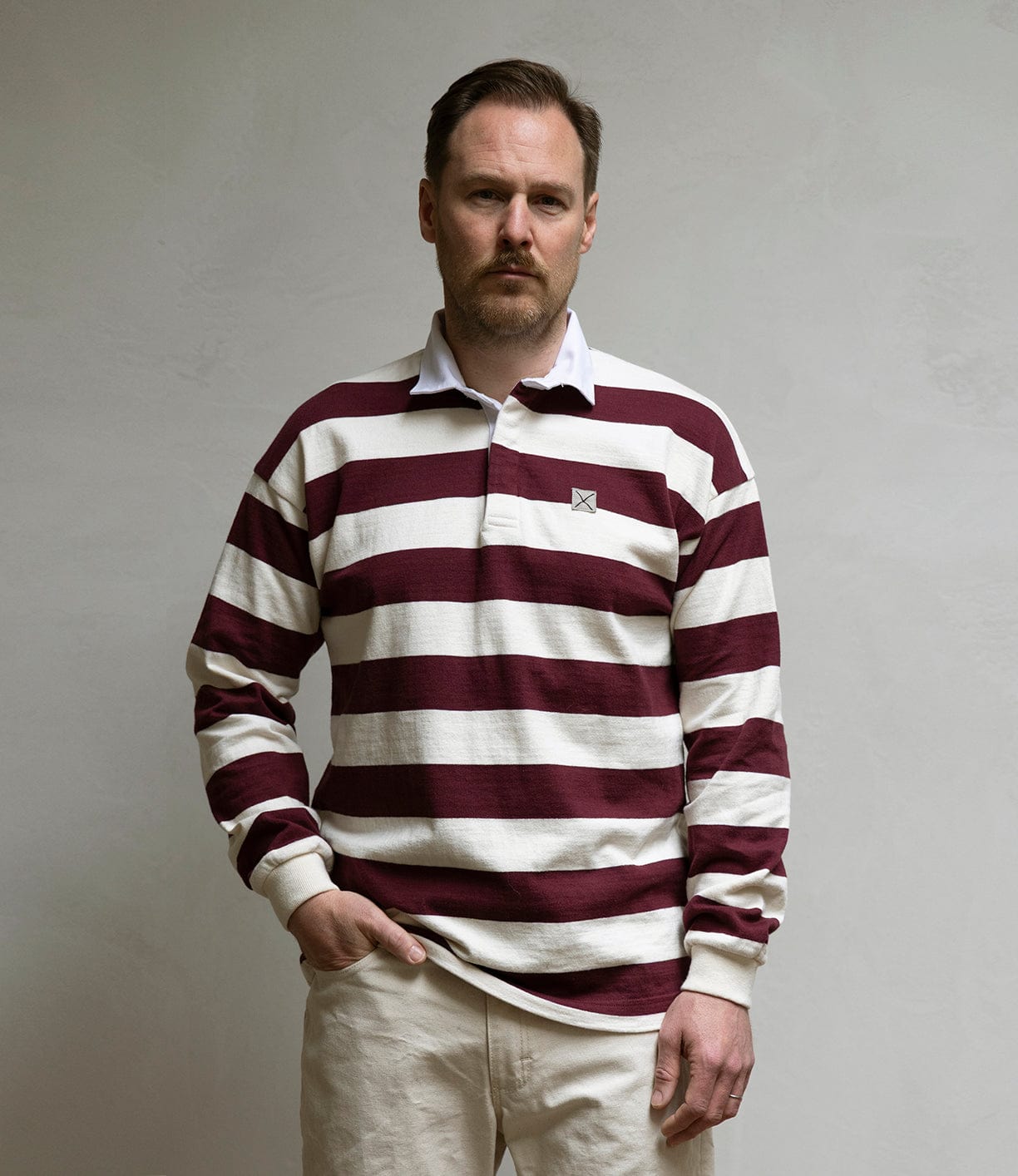 A man with short hair and a mustache wears the Best Made Co MacMillan Rugby Jersey—maroon and white striped heavyweight cotton—standing against a plain light-colored wall, neutral-faced. Made in USA.