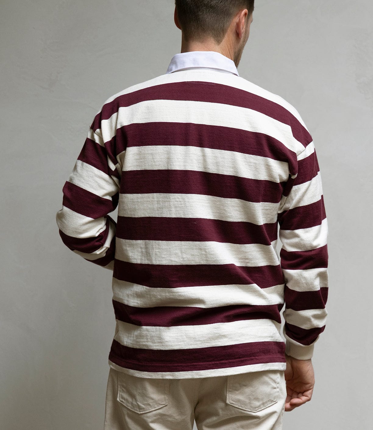 A man, seen from behind, wears the Best Made Co MacMillan Rugby Jersey in maroon and white heavyweight cotton with beige pants, standing before a plain light-colored wall.