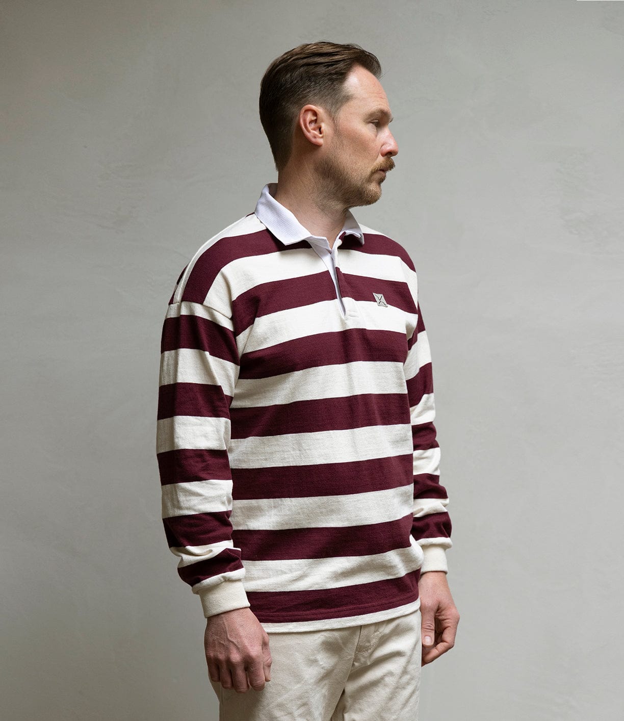 A man stands sideways by a plain light wall, wearing the Best Made Co MacMillan Rugby Jersey—a maroon and white striped heavyweight cotton shirt with a white collar—and light-colored pants.