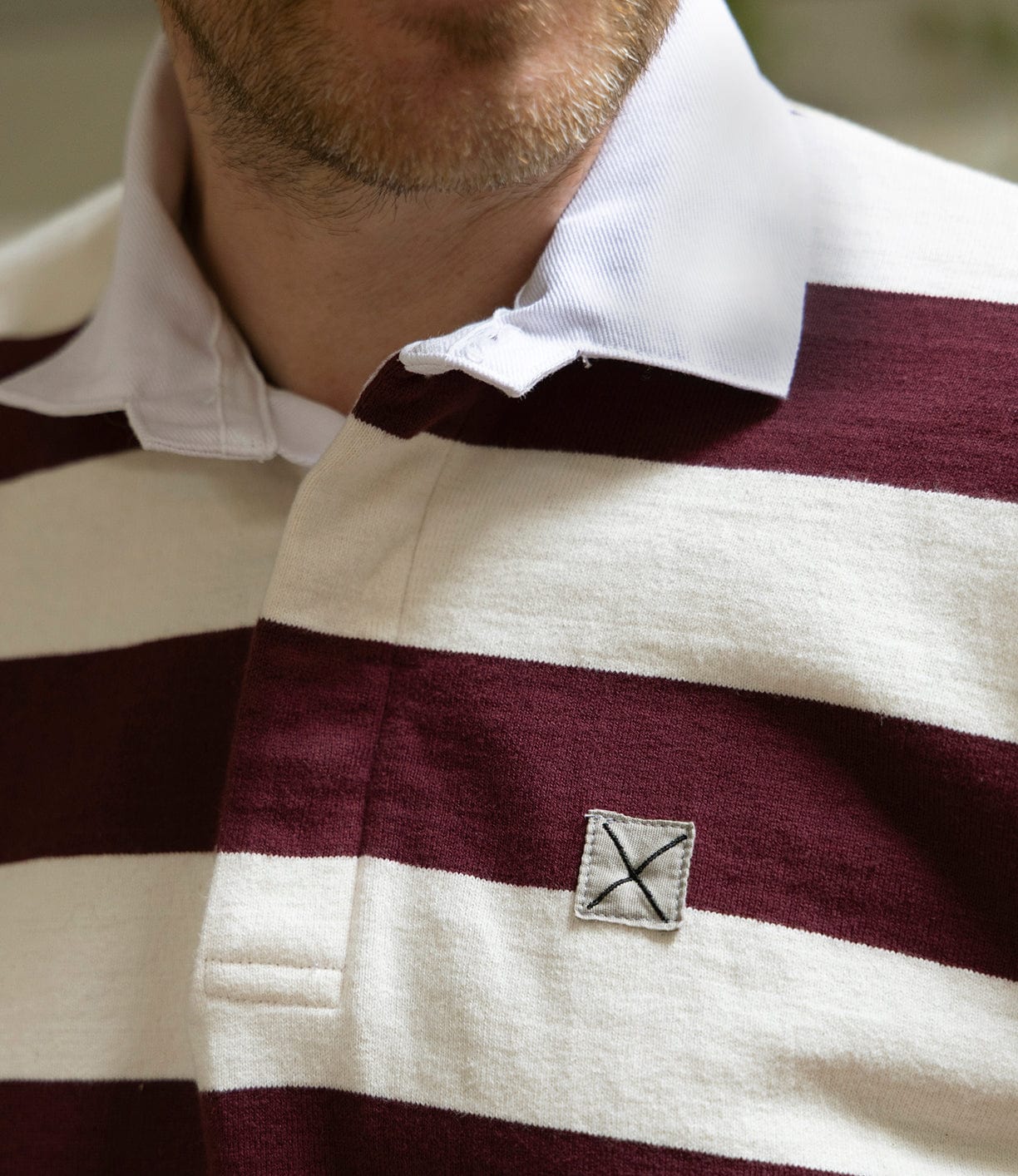 Close-up of a person wearing the Best Made Co MacMillan Rugby Jersey—a maroon and white striped heavyweight cotton shirt with a white collar and small stitched X patch on the chest. Only the lower face and shirt are visible.