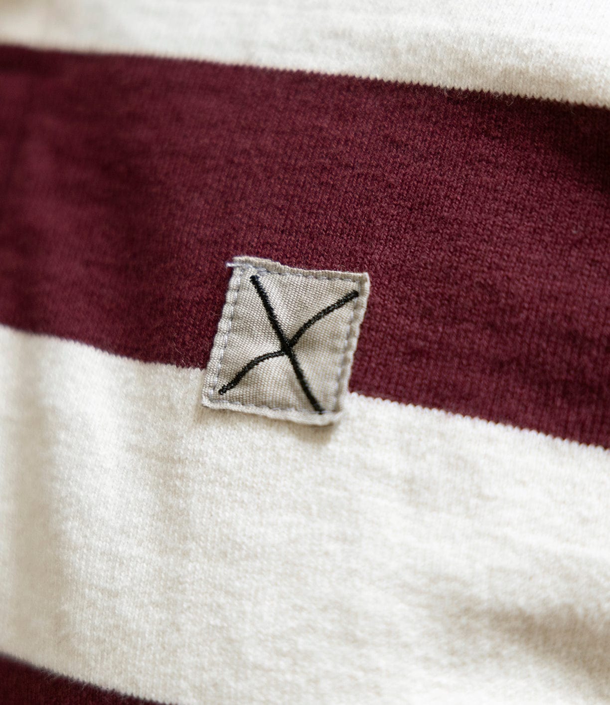 A close-up of the Best Made Co MacMillan Rugby Jersey, featuring wide maroon and white stripes on heavyweight cotton, made in the USA, with a small gray patch sewn on using a black X stitch.
