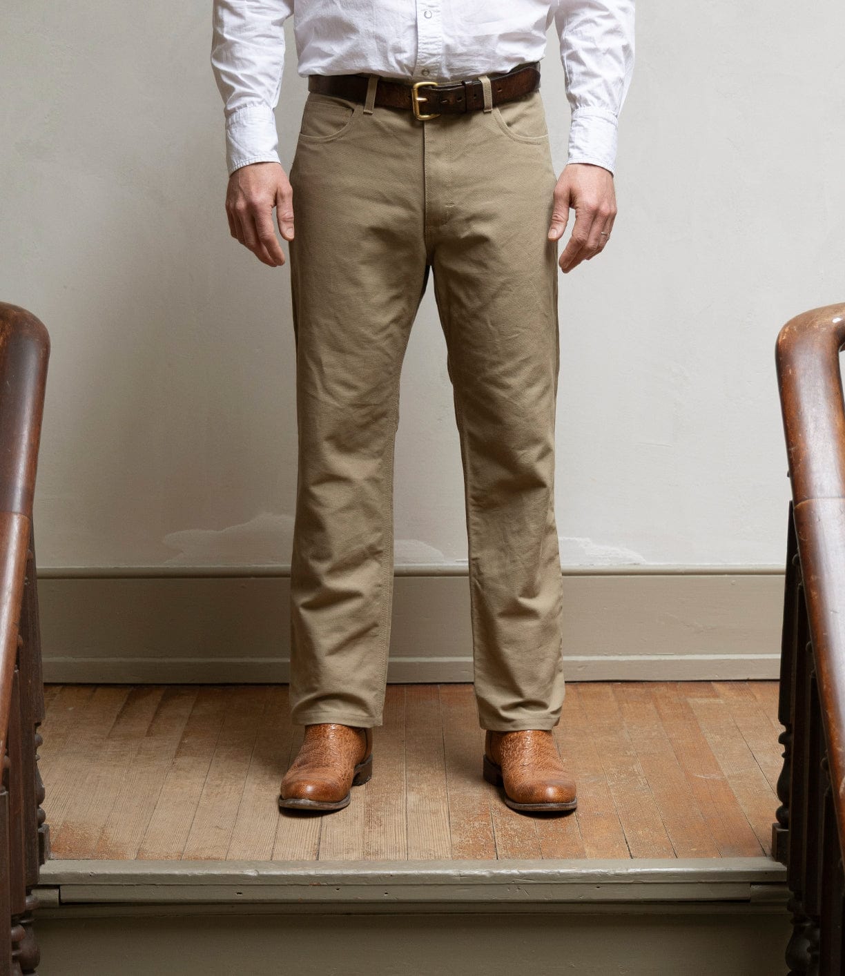 A person stands on a wooden floor, wearing Best Made Co's Canvas 5-Pocket Pant Mark 2 - Undyed, paired with a white button-down shirt, dark belt, and brown boots. The person's head is not shown.