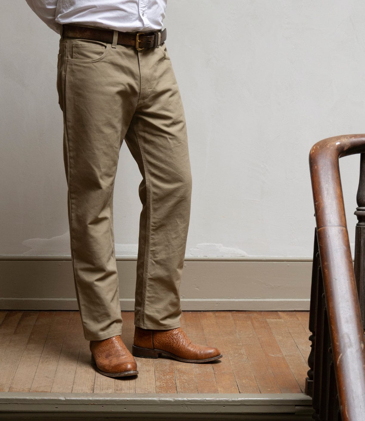 Standing indoors by a staircase on a wooden floor, the individual wears Best Made Co's Canvas 5-Pocket Pant Mark 2 in Undyed, paired with a white shirt, brown belt, and brown leather boots. Their upper body and face are out of frame.