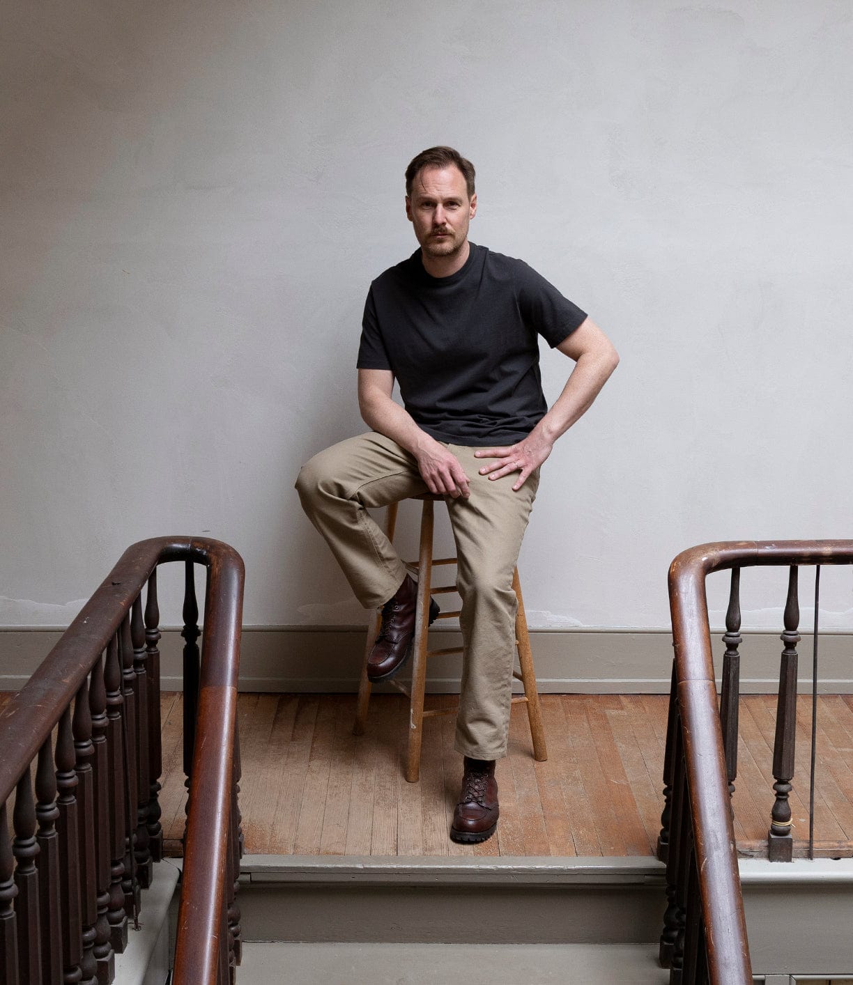 A man with a mustache sits on a wooden stool on a staircase landing, wearing a black t-shirt, brown boots, and the Best Made Co Canvas 5-Pocket Pant Mark 2 - Undyed, against a plain light-colored wall.