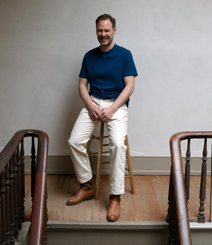 A man with short hair and a mustache, wearing the Best Made Co. Standard T-Shirt in Signal Blue, white pants, and brown boots, sits on a wooden stool in a hallway with wooden floors and a light-colored wall.