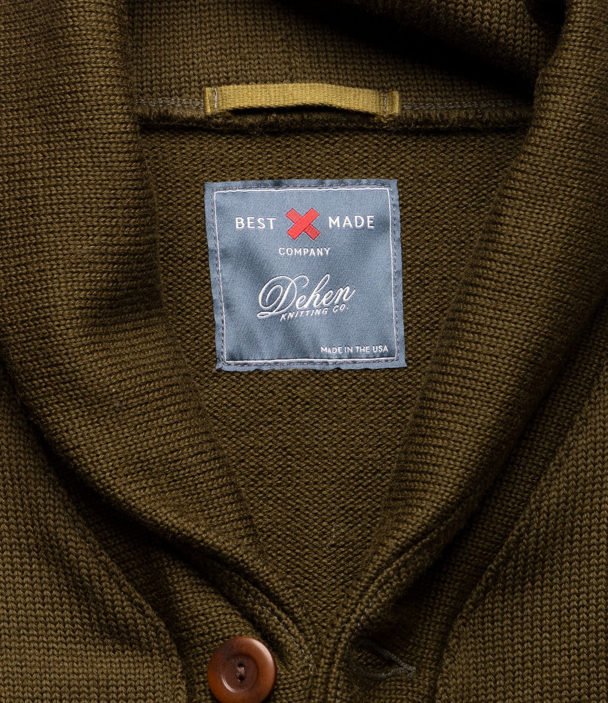 Close-up of the moss green Dehen Shawl Neck Sweater by Best Made Co., featuring textured knit, brown buttons, a shawl neck, and a label sewn below the collar.