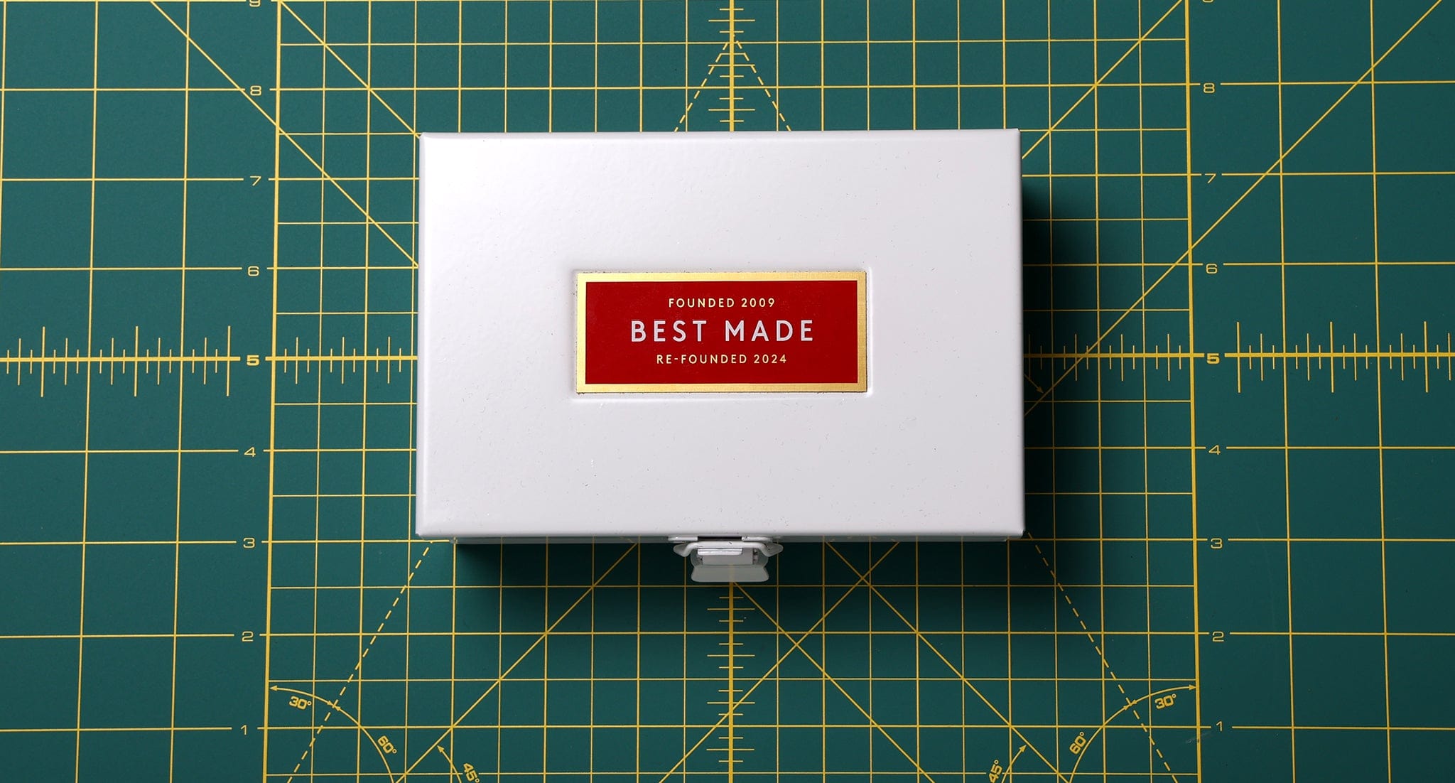 A white metal box with a red and gold label reading Best Made Founded 2009 Re-Founded 2024 sits centered on a green gridded cutting mat marked with yellow measurement lines.