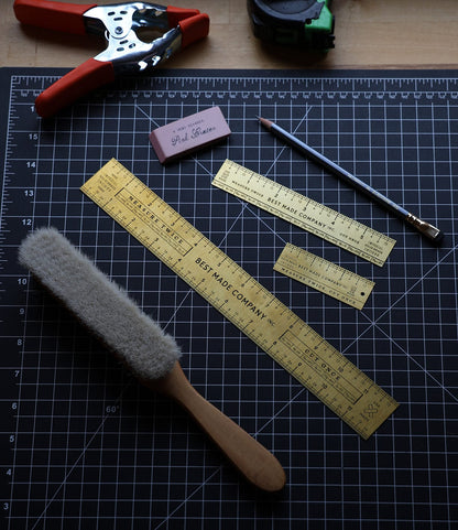 A black grid cutting mat displays the Best Made Co. Measure Twice 12 inch Solid Brass Ruler, precision measuring tools, a pencil, pink eraser, wool duster, tape measure, and a metal hole puncher neatly arranged on top.