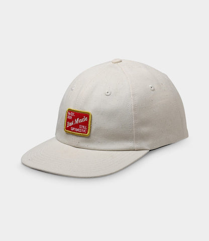 The Selvedge Natural Denim Ball Cap by Best Made Co. is cream-colored with a curved brim, a red and yellow "Best Mode Still Optimistic" patch, and a leather adjustment strap at the back.