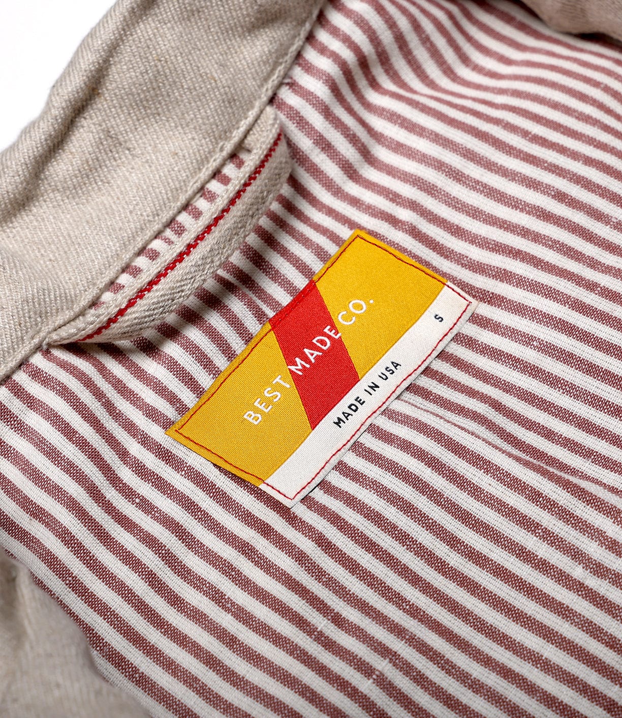 A close-up of the Selvedge Beige Studio Jacket collar by Best Made Co shows red and white vertical stripes, a yellow "BEST MADE CO." tag, and precise stitching—details that showcase American craftsmanship.