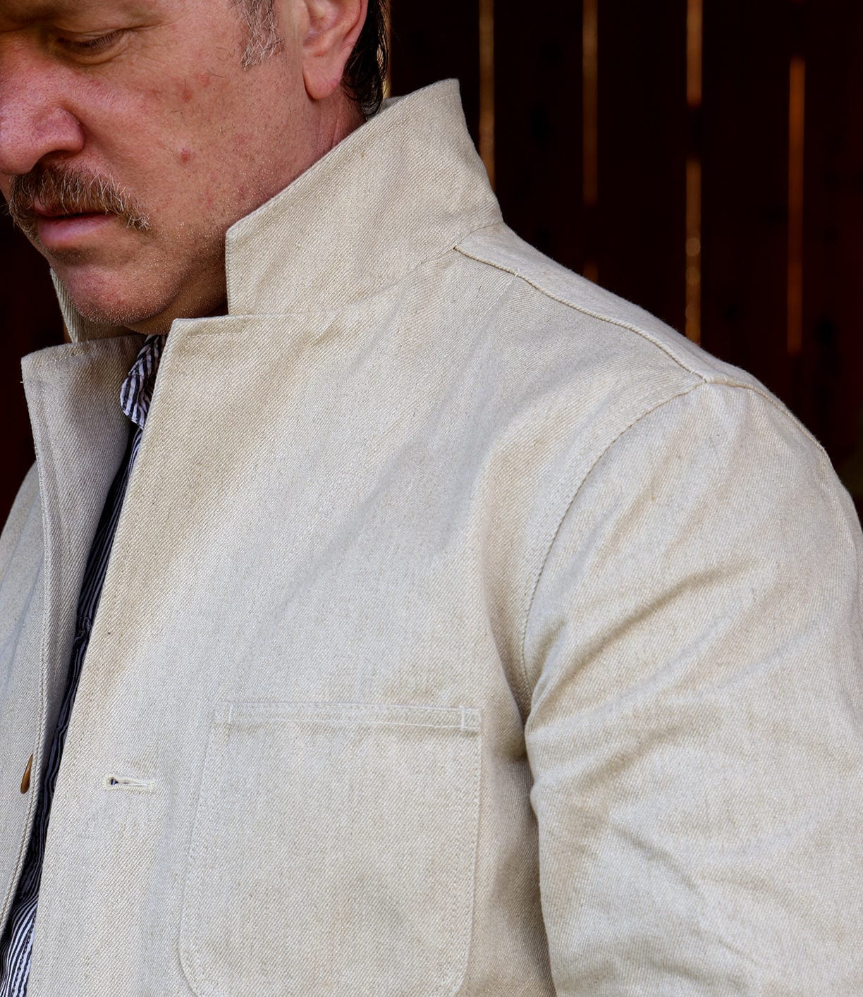 A mustached man stands against a dark background, showcasing the Best Made Co Selvedge Beige Studio Jacket with a high collar and chest pocket; only his lower face and upper torso are visible, highlighting American craftsmanship.