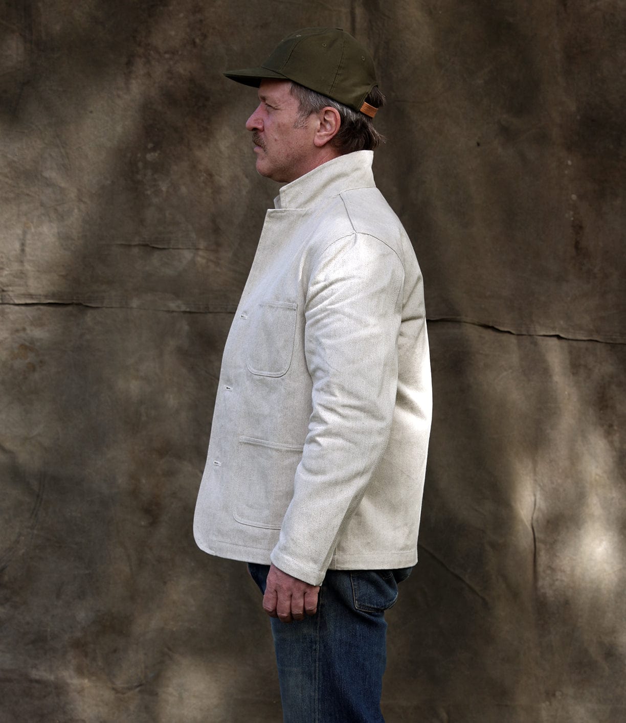 A man in a Best Made Co Selvedge Beige Studio Jacket, blue jeans, and an olive cap stands in profile against a textured brown backdrop, exemplifying American craftsmanship.