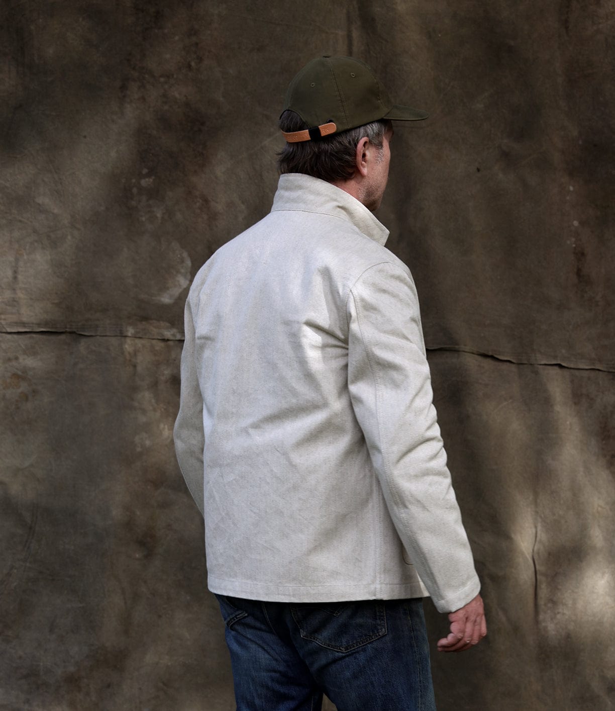 Wearing a Best Made Co Selvedge Beige Studio Jacket, green cap, and blue jeans, a person stands with their back to the camera against a textured brown backdrop, exemplifying American craftsmanship.