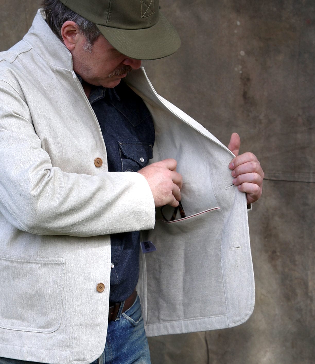 A man in a Best Made Co Selvedge Beige Studio Jacket, blue shirt, and green cap opens his jacket to access an inner pocket, standing before a plain brown background—a tribute to American craftsmanship.