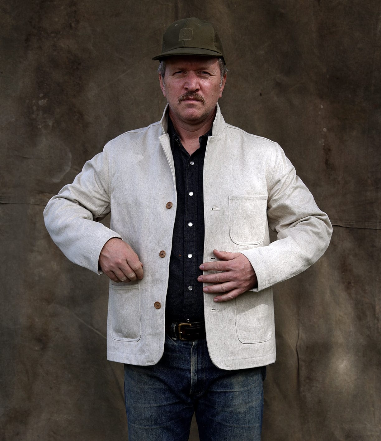A man wearing the Selvedge Beige Studio Jacket by Best Made Co, a dark shirt, blue jeans, and an olive green cap stands against a brown textured background, looking forward with a neutral expression that highlights American craftsmanship.