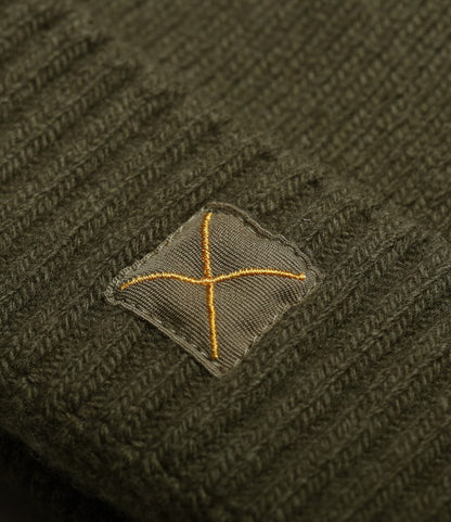 A close-up of a dark green knitted fabric, likely the Best Made Co. Cap of Courage, featuring a stitched square patch with a yellow X in the center.