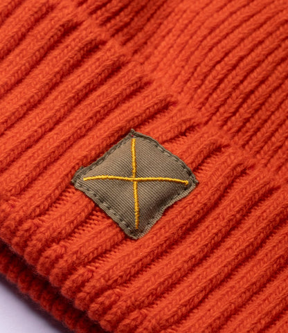 Close-up of an orange ribbed knit Cap of Courage by Best Made Co. featuring a small olive green square patch with a yellow stitched X design.
