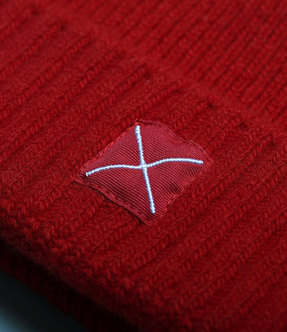 Close-up of the Cap of Courage by Best Made Co., showing red knit lambswool fabric and a stitched patch with a white “X,” highlighting both the cap’s texture and detailed patchwork.