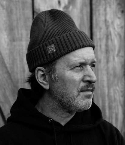 Black and white photo of a bearded, middle-aged man in a Best Made Co. Cap of Courage knit beanie and hoodie, gazing thoughtfully to the side with a wooden wall in the background.