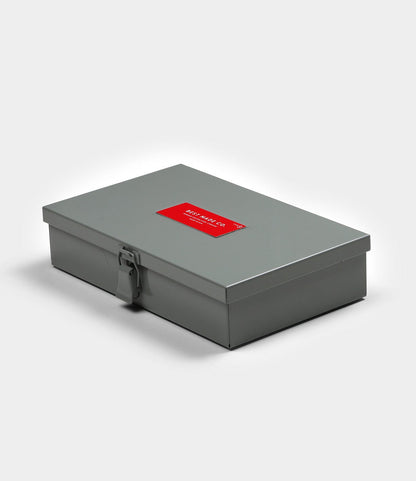 The Best Made Co. Type 9 Strongbox is a closed, rectangular gray steel storage box featuring a front latch and a red label with white text on the lid, shown on a plain white background.
