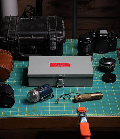 A tabletop with a green cutting mat displays several items: a Best Made Co. Type 9 Strongbox with a red label, a sturdy black case, a Nikon camera with lenses, a flashlight, a brass tool, a clamp, and a brown leather case.