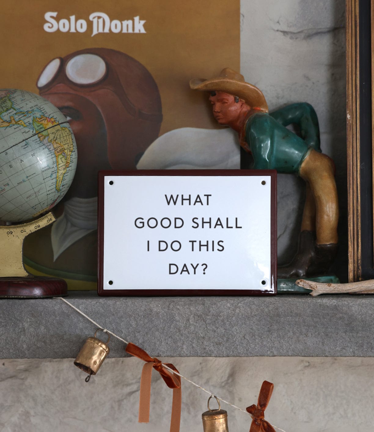 A shelf displays a vintage globe, a cowboy figure, a framed “Solo Monk” album, and Best Made Company Co.'s "Stay Sharp" Enamel Steel Sign (kanji edition). A string with bells hangs below, offering an inspiring touch.