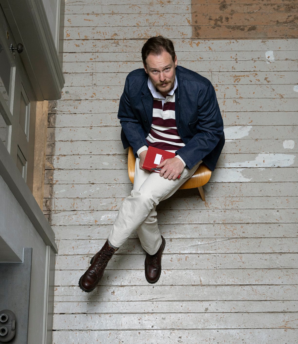 A bearded man in a navy jacket, light pants, and brown boots sits on a worn wooden floor holding a red notebook, wearing the Best Made Co MacMillan Rugby Jersey in maroon and white stripes, looking up at the camera.