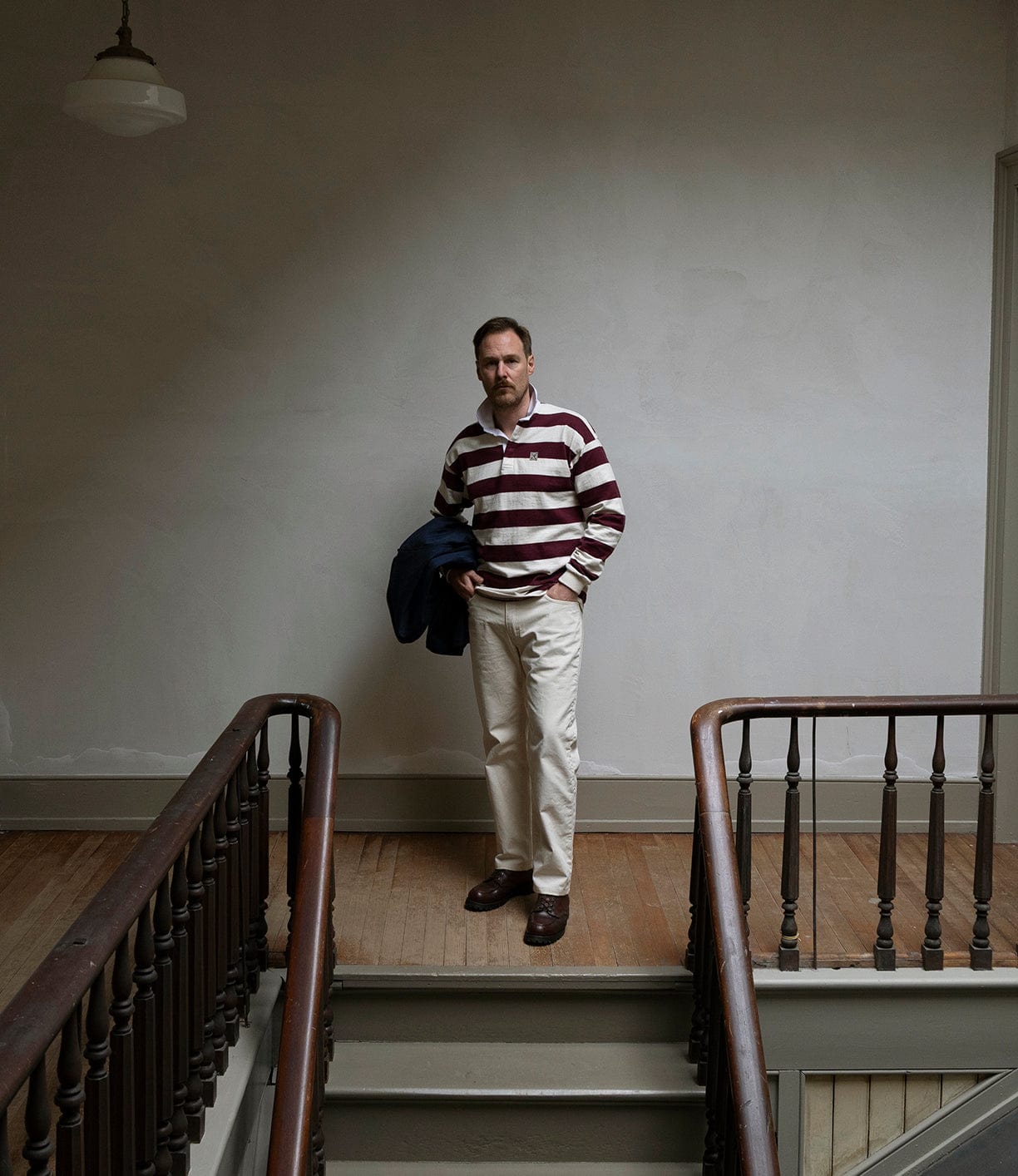 A man stands on a landing between two wooden staircases, holding a jacket. He wears the Best Made Co MacMillan Rugby Jersey—maroon and white stripes, made in the USA—paired with beige pants and brown shoes. The wall behind him is plain and pale.