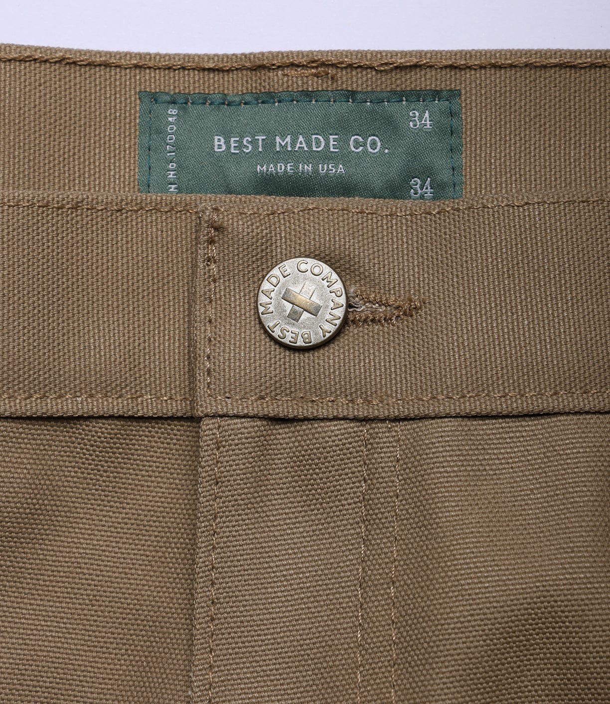 Close-up of the Best Made Co Canvas 5-Pocket Pant Mark 2 - Undyed, featuring a metal button engraved with “BEST MADE COMPANY” and a green waist label marked “BEST MADE CO. 34 MADE IN USA,” size 34 shown on both label and waistband.