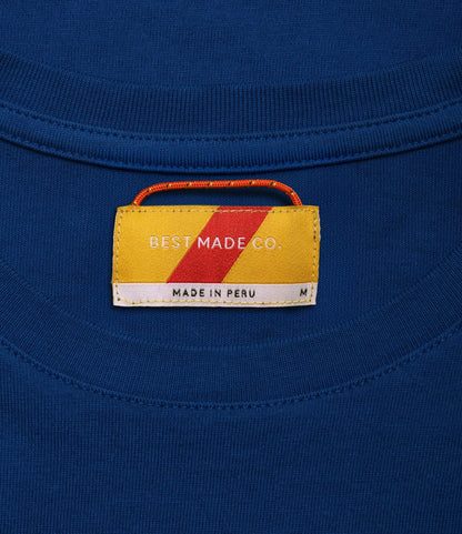 A close-up of the Standard T-Shirt Signal Blue by Best Made Co. shows a yellow and red label with "BEST MADE CO." and "MADE IN PERU," plus an M for medium—crafted from durable Peruvian tangüis cotton.