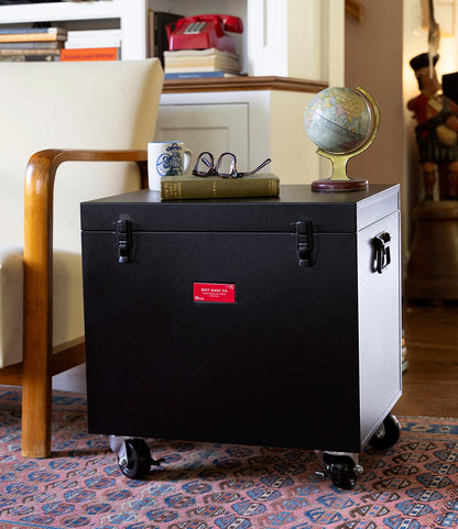 The Best Made Co. Rolling Footlocker, used as a side table, holds a book, eyeglasses, mug, and small globe beside a wooden armchair on a patterned rug in a cozy room—offering stylish storage and decor.