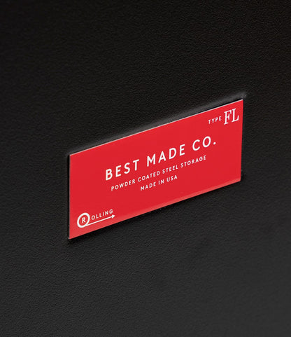 A red label on a black, 22-gauge steel Rolling Footlocker by Best Made Co. reads: BEST MADE CO. POWDER COATED STEEL STORAGE MADE IN USA TYPE FL, with a rolling logo in the bottom left—a durable storage solution.
