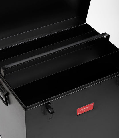 The Best Made Co. Rolling Footlocker is a black 22-gauge steel storage box with an open lid, sturdy handle, inner compartment, front latch, and a small red label—an ideal storage solution for any space.