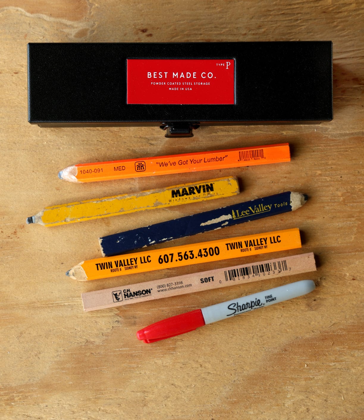 A Best Made Co. Type P Strongbox, Go Slow Edition, rests on a wooden surface above six assorted carpenter pencils and a gray Sharpie marker with a red cap, all neatly arranged.