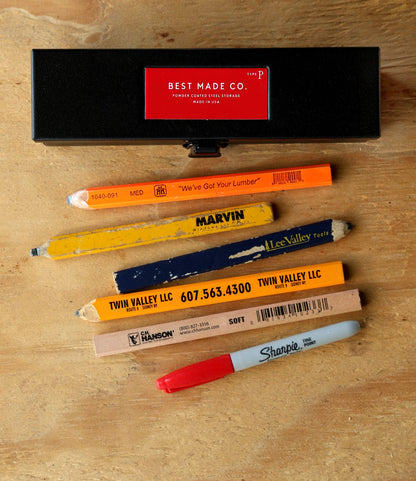 A Best Made Co. Type P Strongbox in matte black sits above five assorted carpenter pencils and a red-and-black Sharpie, all arranged on a wooden surface. The pencils display various brand names and colors.