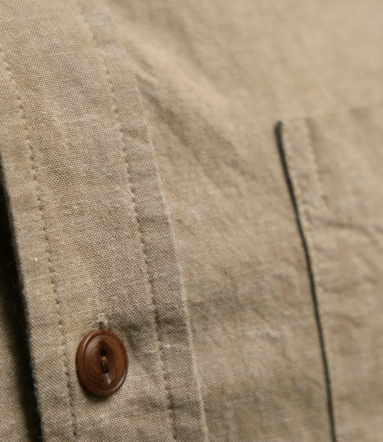 Close-up of the Best Made Co. Cotton/Linen Herringbone Workshirt in Tan, featuring a brown button and part of a pocket. The fabric's textured, woven look echoes classic Japanese chambray.