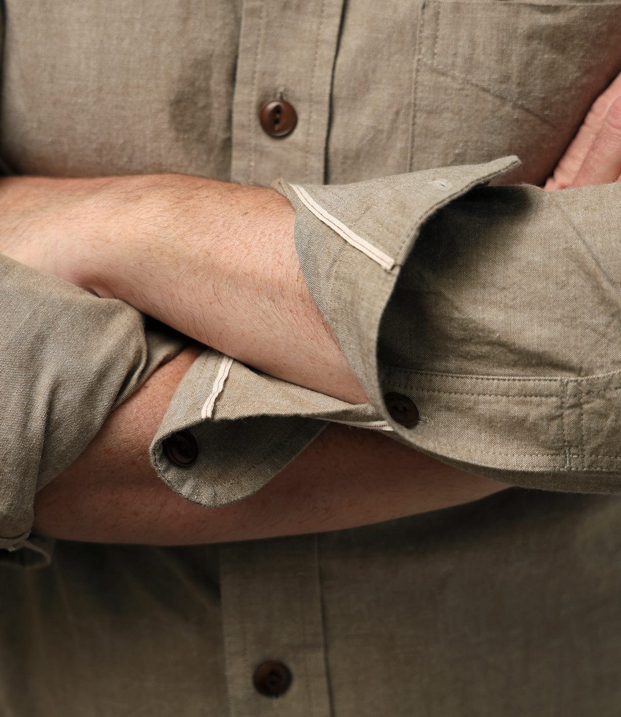 A person wearing the Best Made Co. Cotton/Linen Herringbone Workshirt in Tan stands with arms crossed and sleeves partially rolled up, showing only their torso and arms.