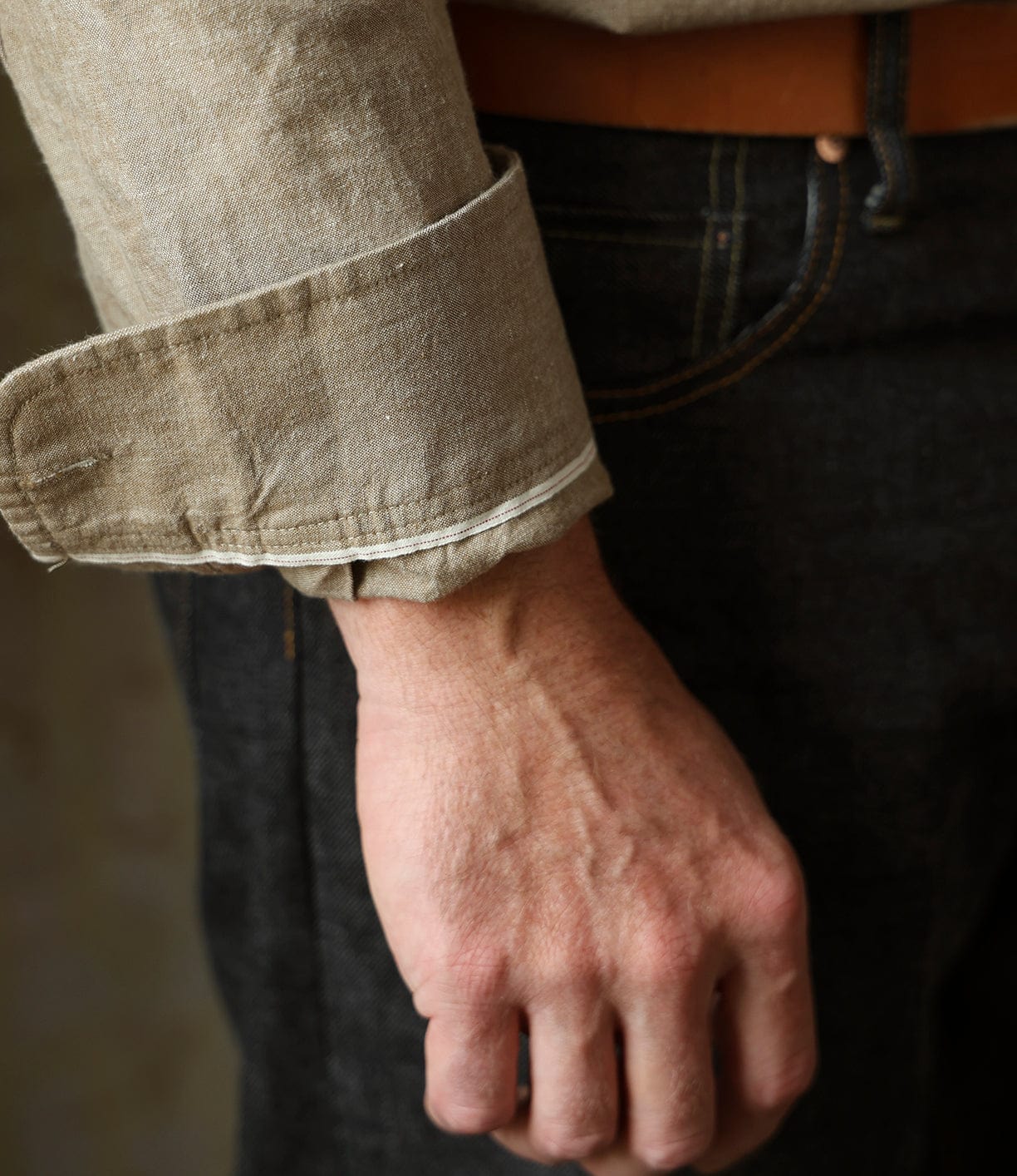 A close-up of a person’s hand and wrist wearing the Best Made Co. Cotton/Linen Herringbone Workshirt in Tan, with the sleeve rolled up, paired with dark denim jeans and a brown belt. The relaxed hand is slightly clenched.