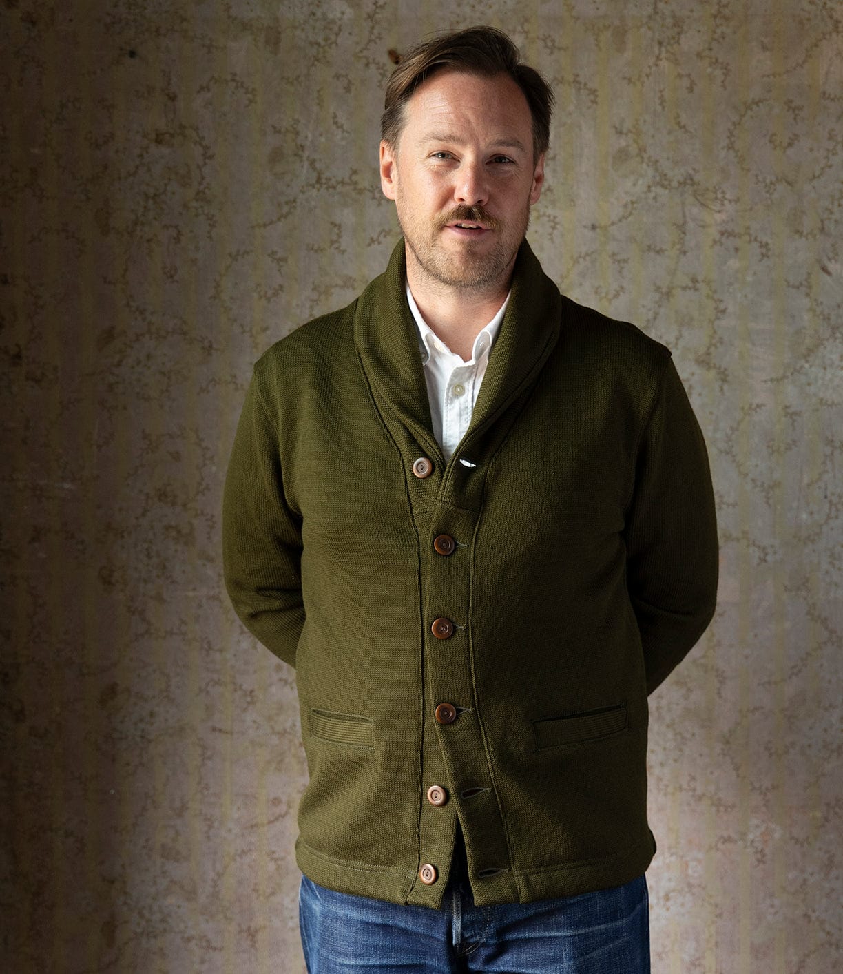 A man with light brown hair and a trimmed beard stands against a textured beige wall, wearing the Best Made Co. Dehen Shawl Neck Sweater in Moss Green over a white shirt and blue jeans, his hands behind his back.