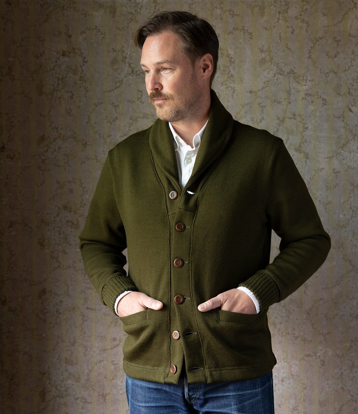 A man with short brown hair and a beard stands against a patterned wall, wearing the Best Made Co. Dehen Shawl Neck Sweater in Moss Green over a white shirt and blue jeans, his hands in his pockets as he looks to the side.