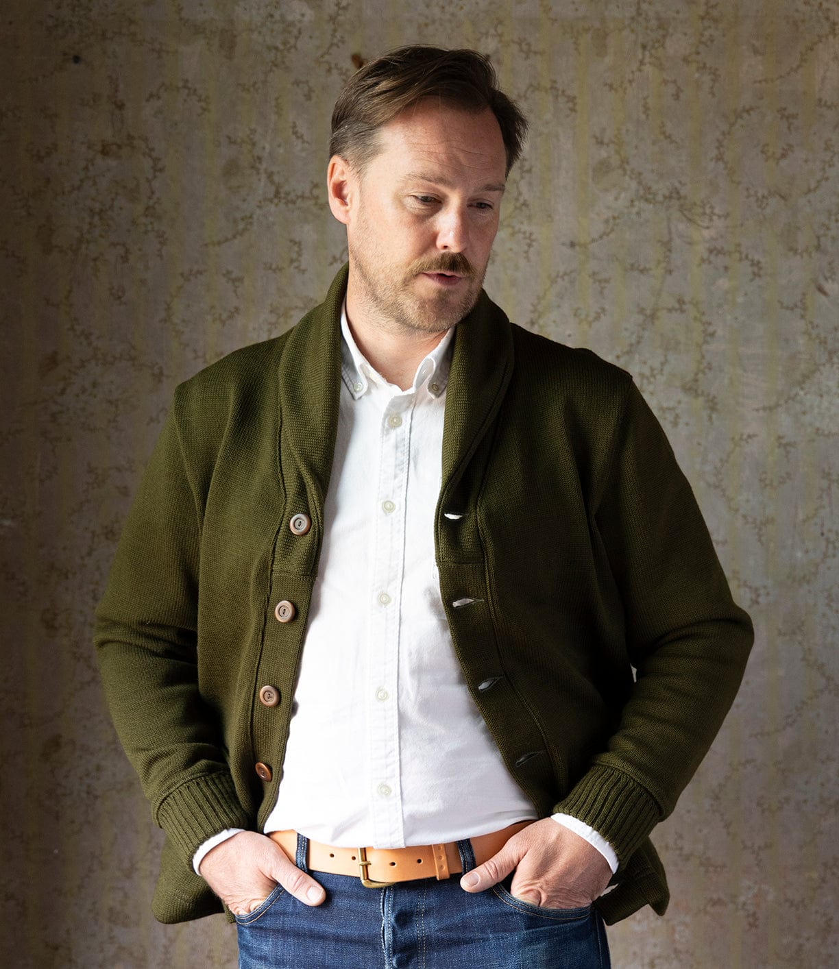 A man with light brown hair and a mustache stands against a beige wall, wearing a white shirt, blue jeans, tan belt, and the Best Made Co. Dehen Shawl Neck Sweater in Moss Green—channeling classic American workwear—hands in pockets, gazing downward.